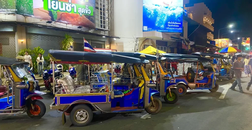 A line of traditional blue tuk-tuks waiting at night near the festival gates with neon signs and YUU Hotel in the background.