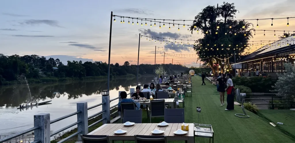 Sunset dining terrace with warm ambient light strands and organized wooden tables along the quiet Mun River in Ubon Ratchathani, Isaan.