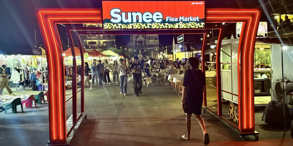 The accessible concrete entrance under a glowing neon arch at Sunee Night Market in Ubon Ratchathani, showing local clothing stalls and an open-air dining area.